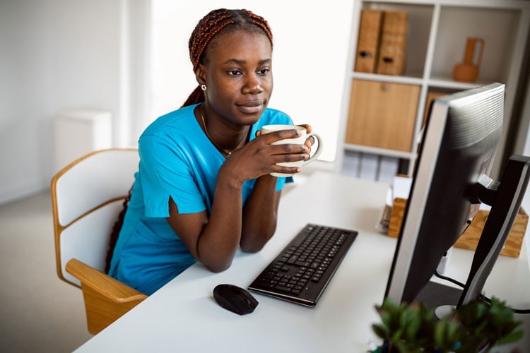 Caregiver in blue scrubs drinking coffee while working on a computer at home.